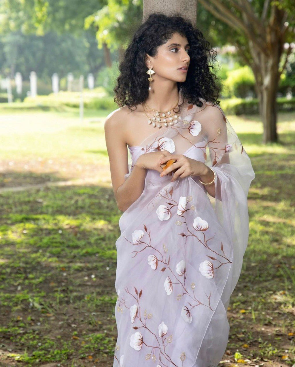A model wearing the sheer lavender floral print organza saree with delicate silver handwork, paired with a matching blouse, standing gracefully in a park.