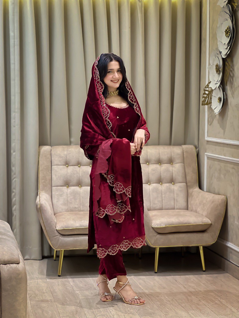 Model smiling in a full-length shot, wearing a maroon velvet salwar suit set with intricate rhinestone work, standing in an elegant room.