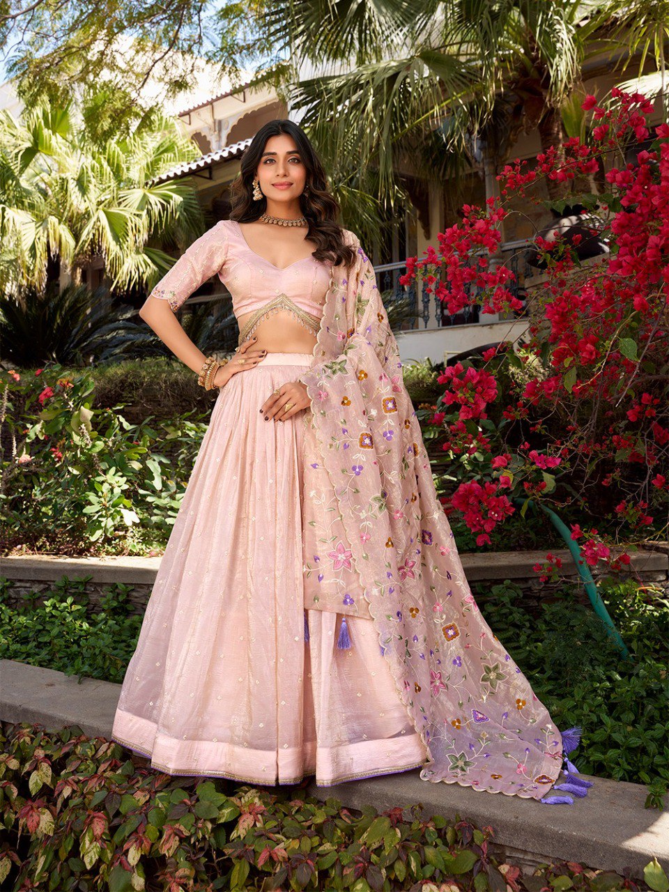 Woman posing in a blush pink Gold Crush lehenga choli with sequin and floral embroidered dupatta in a lush garden, full view.