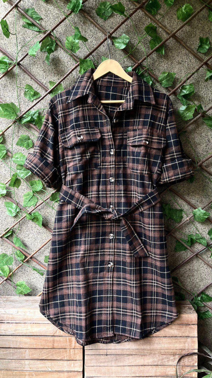 TRENDBUY brown plaid flannel shirt dress with belt on a hanger, styled against a rustic wall with green foliage.