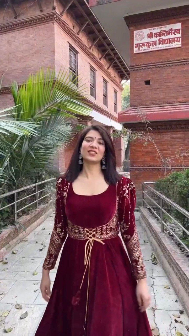 Woman gracefully posing in the red velvet Anarkali gown and sequin jacket against a historical building.