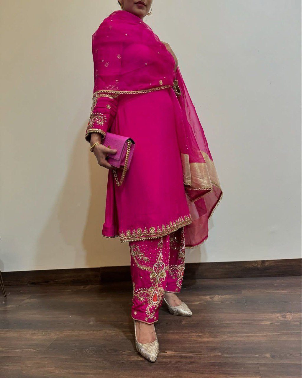 Model posing in a radiant fuchsia pink faux georgette palazzo suit with intricate gold embroidery and pearl details, holding a matching clutch.