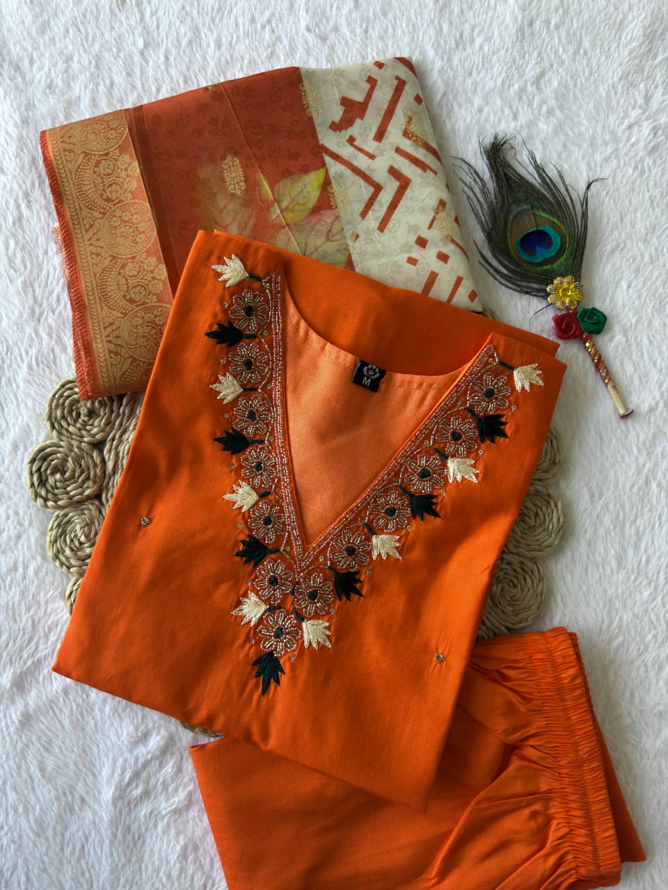 Close-up of Orange Handwork Kurti Pant Dupatta Set, folded neatly on a woven mat with peacock feather.
