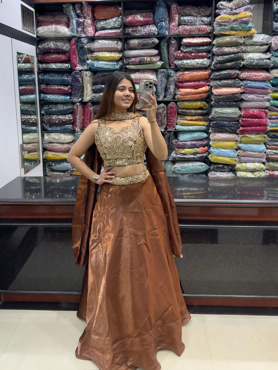 Woman in designer heavy faux lehenga choli set with gold embroidered crop top and bronze skirt posing in front of fabric display.