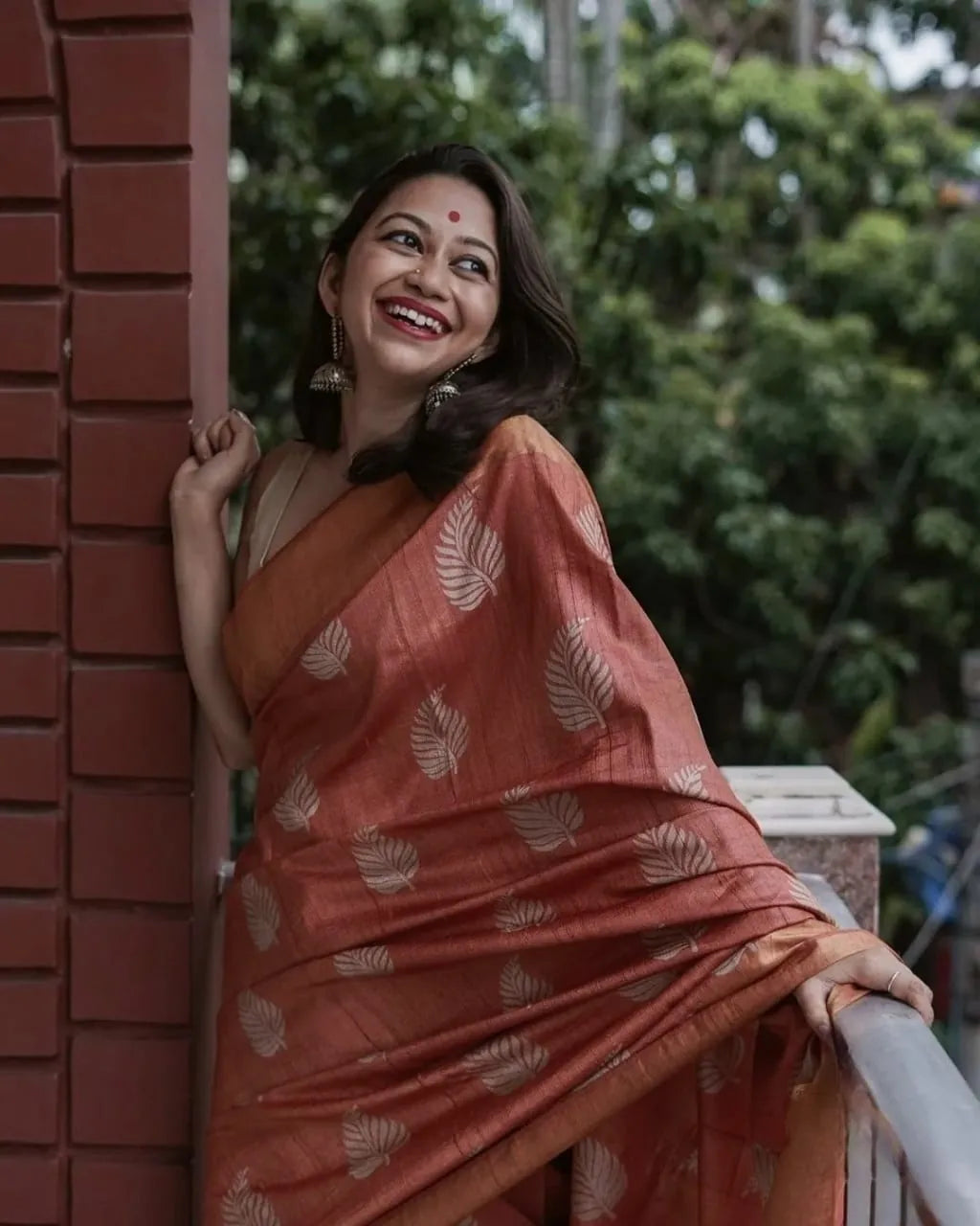Woman wearing rust red banarasi soft silk saree leaning against red brick pillar