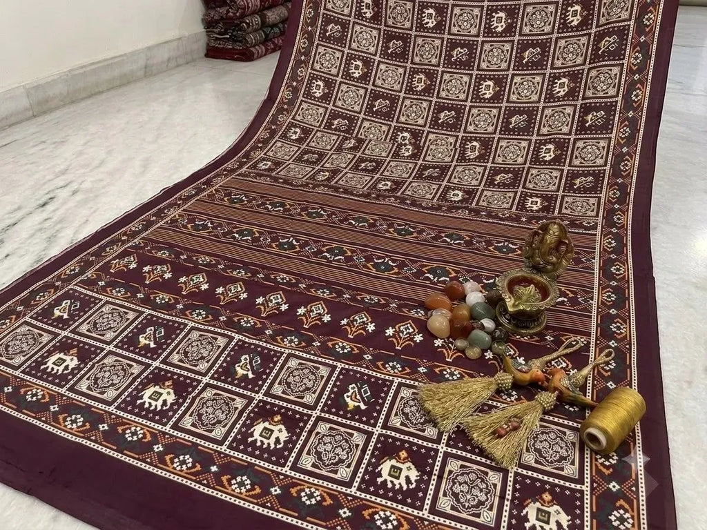 Maroon rayon patola print saree laid flat with decorative Ganesha idol and tassels