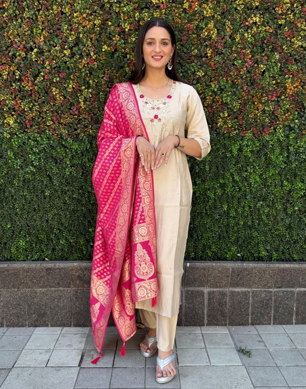 Woman smiling in full length, wearing cream Roman silk kurti pant dupatta set with a vibrant pink jacquard dupatta against a green foliage background.