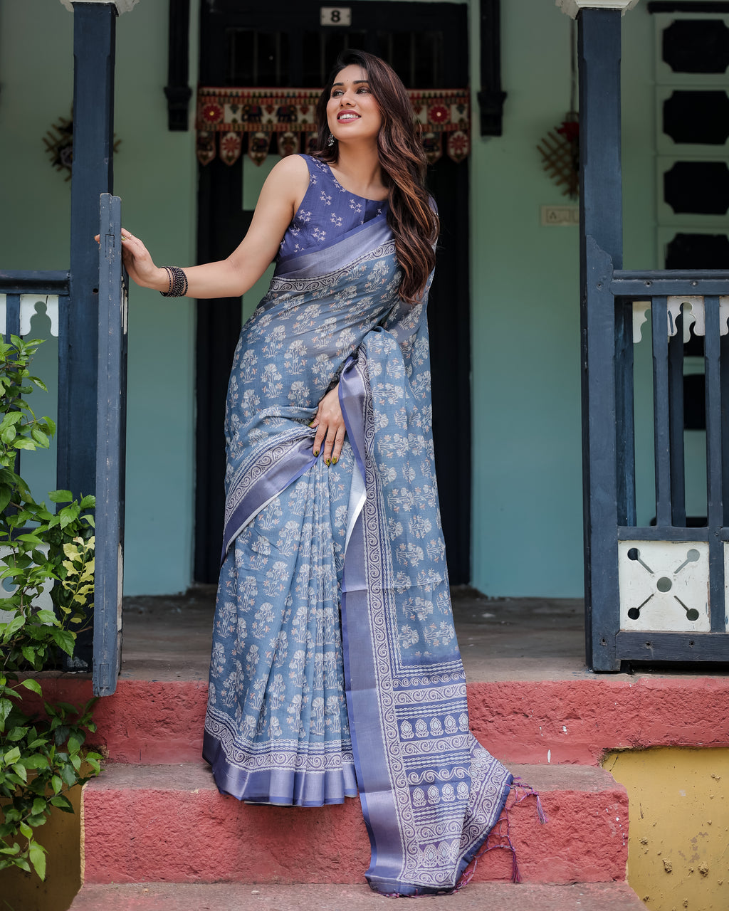 Woman in Gentle Sky Blue Pure Linen Saree with floral prints and tassels, standing by a doorway, full body shot.