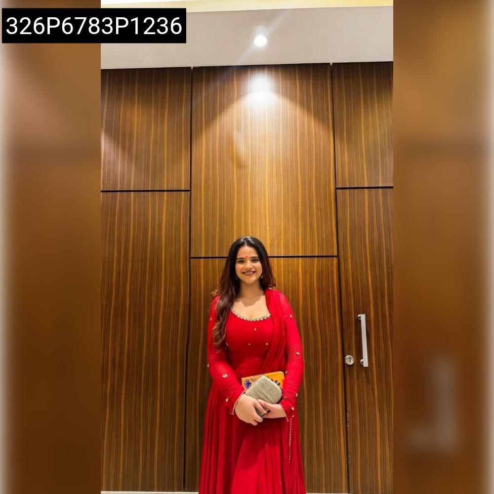Full body shot of model in flowing red Georgette Anarkali suit, holding a clutch, smiling against wooden background.