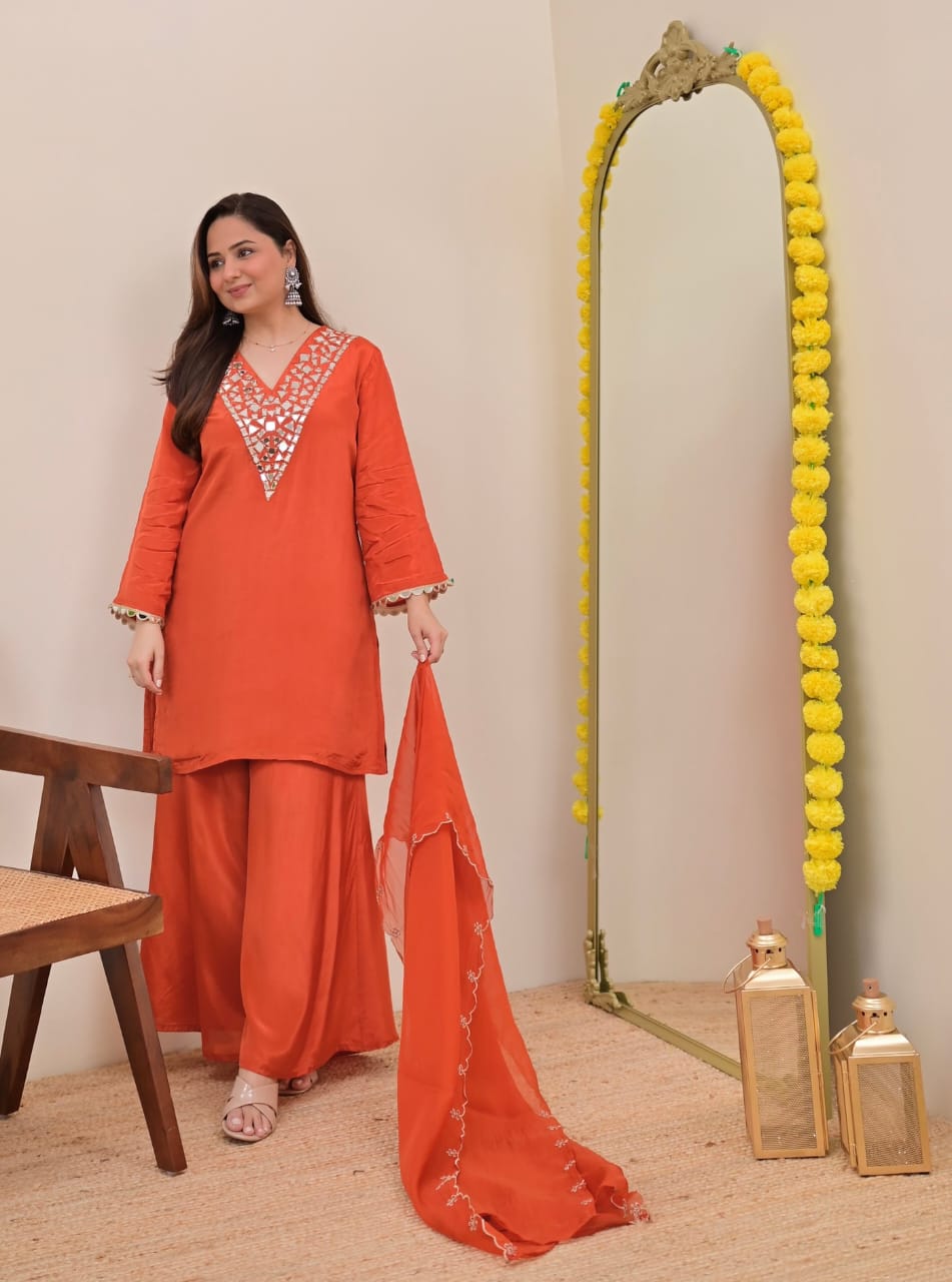 Model in Rust Orange Sharara Set with Mirror Work, standing next to a mirror with marigold garland, showcasing festive attire.