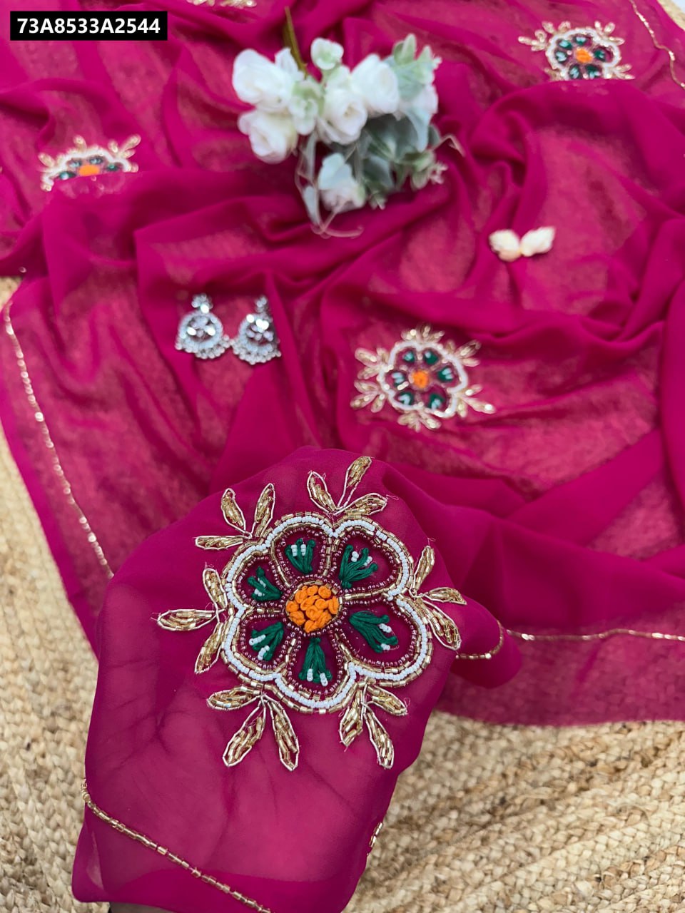 Flat lay of a vibrant hot pink soft Georgette saree with floral moti and thread handwork, styled with earrings and white flowers.