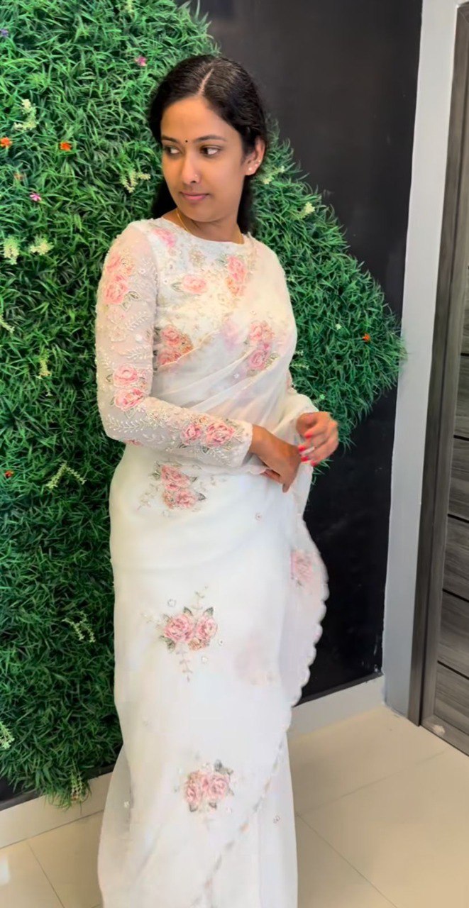 Woman showcasing white Tebi silk saree with pink floral digital print and handwork details, standing against a green wall.