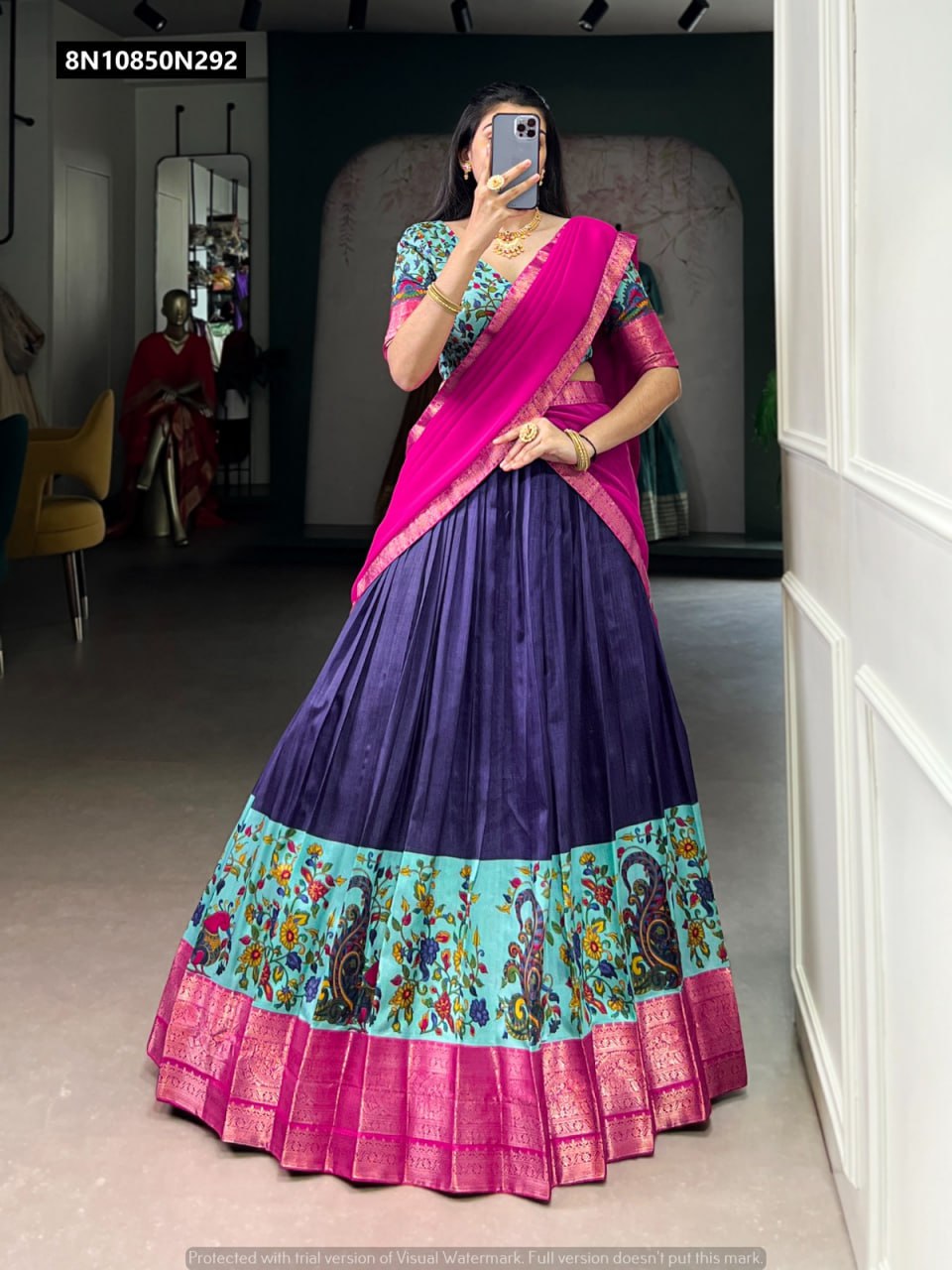 A woman posing elegantly in a mirror, wearing a vibrant TRENDBUY lehenga choli featuring a purple skirt with a printed border, a hot pink dupatta with zari detailing, and a multi-color floral blouse, adorned with traditional jewelry.