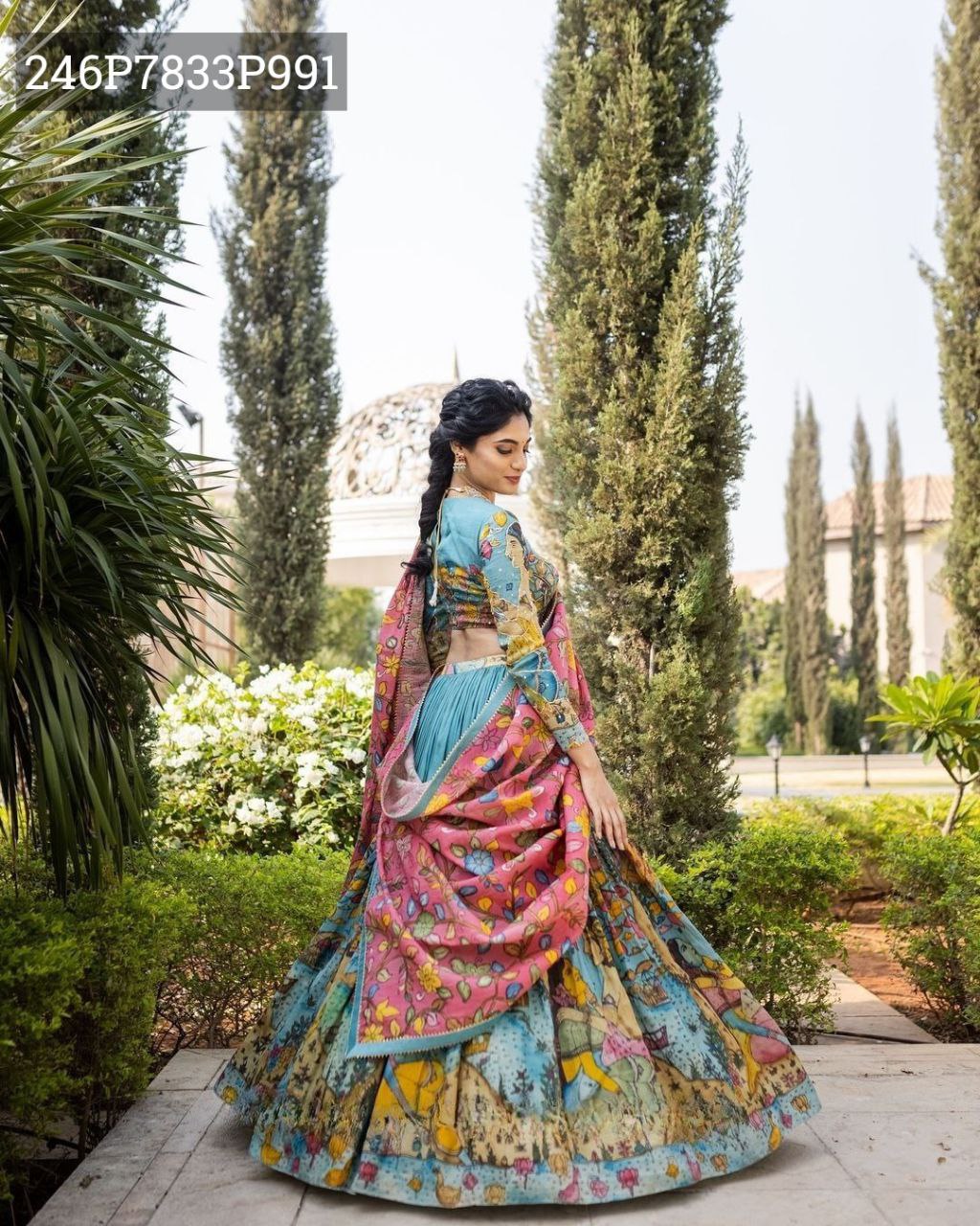 Model posing gracefully in TRENDBUY Aqua Tussar Silk Lehenga Choli with multi-color Kalamkari print and pink floral dupatta, viewed from the back-side in a lush garden setting.