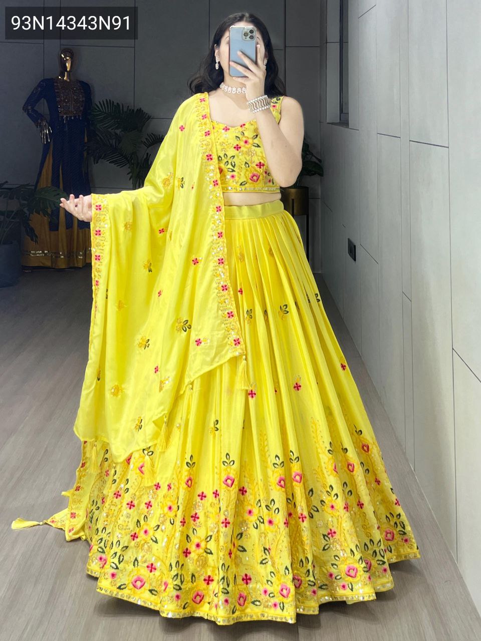 Full body view of a woman showcasing the TRENDBUY classical yellow lehenga choli, featuring a voluminous A-line skirt with multi-color floral embroidery and a matching sleeveless blouse and draped dupatta, standing in a brightly lit hallway.