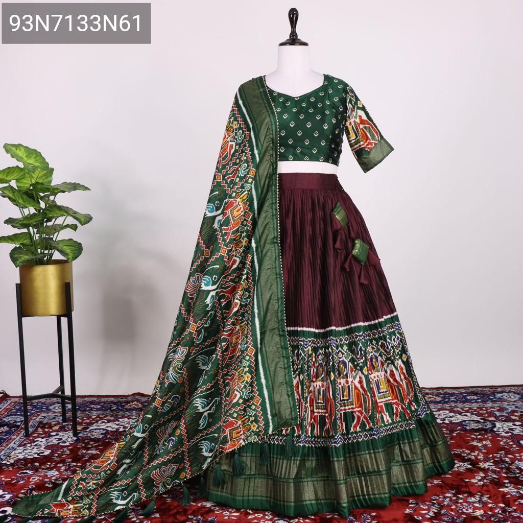 Elegant mannequin displaying the Emerald Green blouse and maroon crushed Dola Silk lehenga with a vibrant Patola print dupatta, set against a subtle background with a plant.