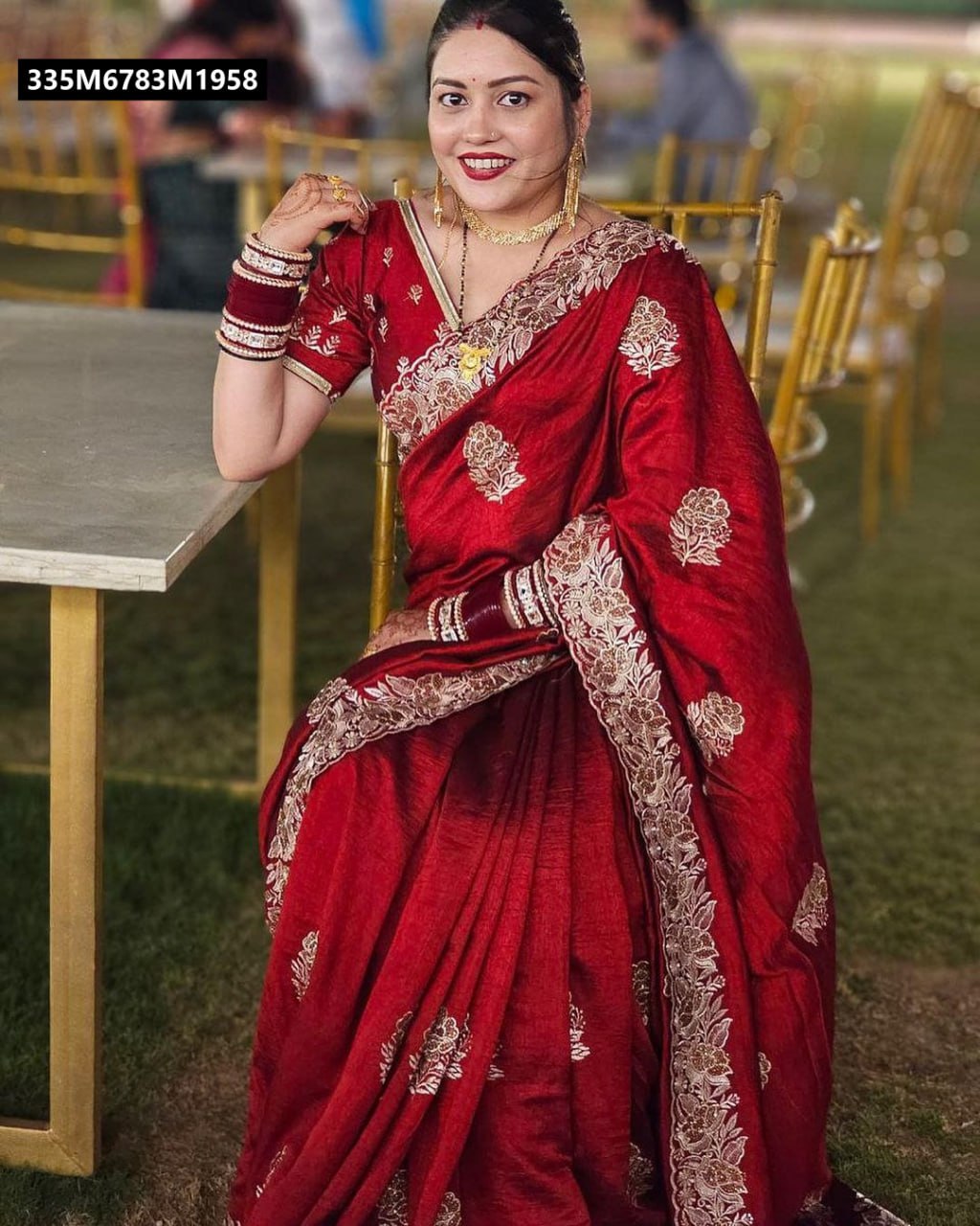 A woman seated at an outdoor event, wearing a radiant red Blooming Vichitra saree with prominent zari and stone work, smiling.