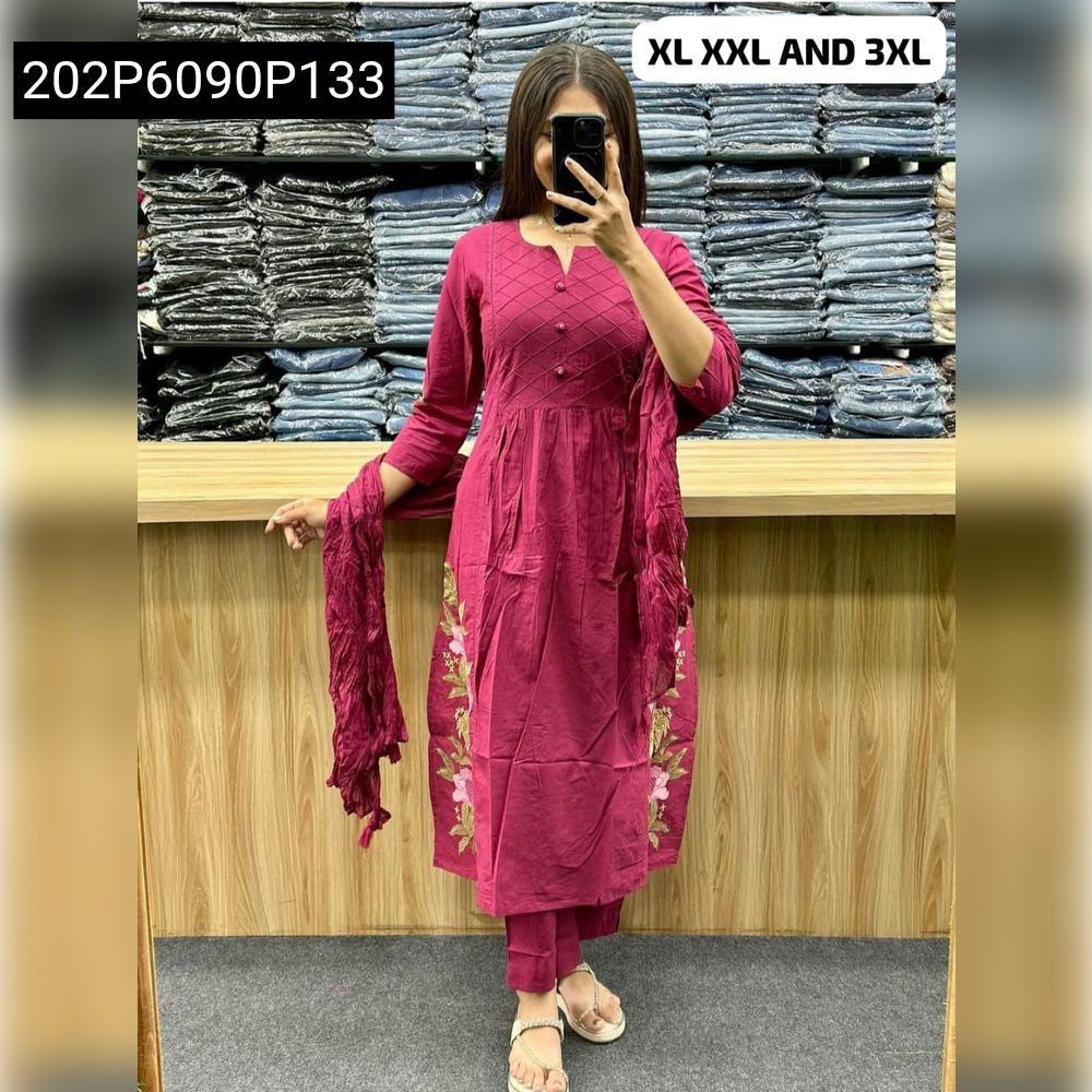 Full body shot of a woman wearing a TRENDBUY magenta cotton flared kurti set with floral embroidery on the hem, matching straight pants, and a crinkled dupatta, posing in front of a clothing rack.