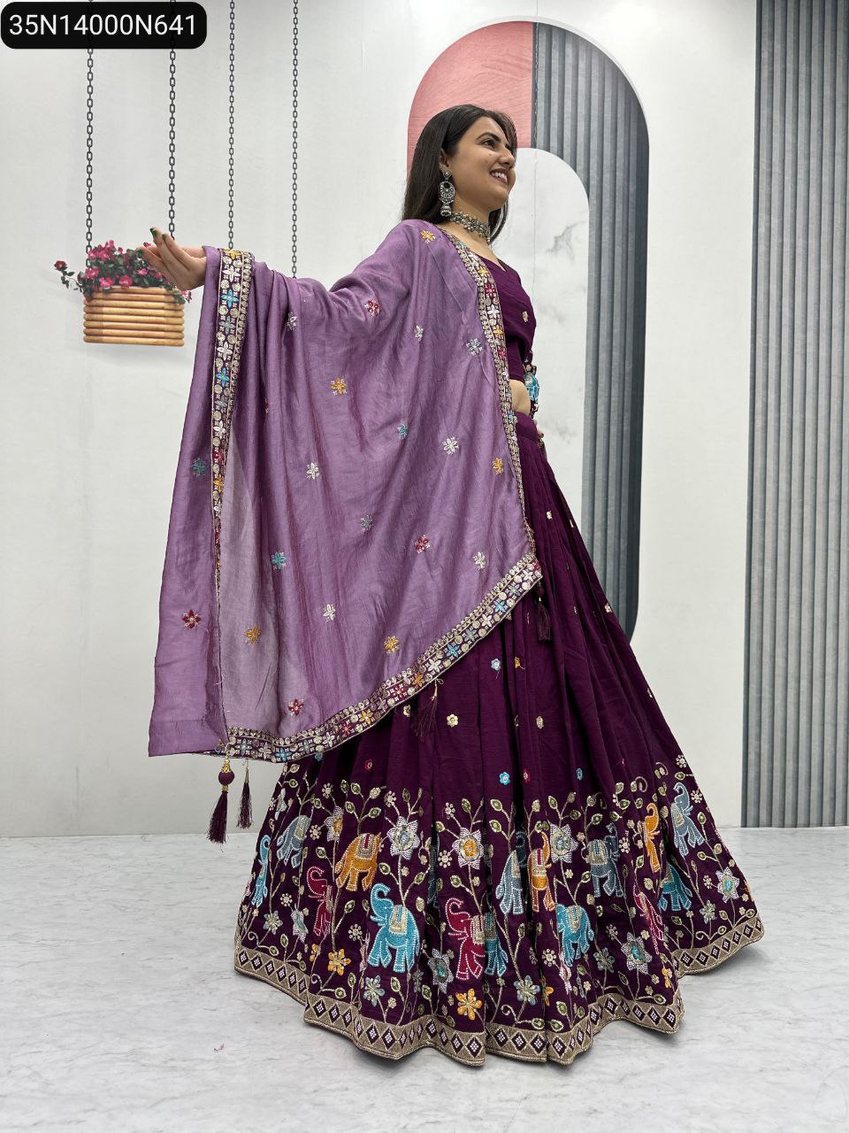 A smiling model wearing a majestic purple Vichitra silk lehenga choli set with vibrant elephant and floral embroidery, holding out her purple dupatta elegantly.