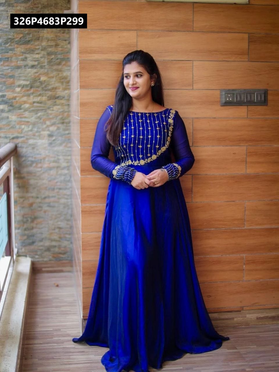 Model wearing elegant Royal Blue Georgette Anarkali Gown with gold leaf and flower embroidery, leaning against a wooden wall.