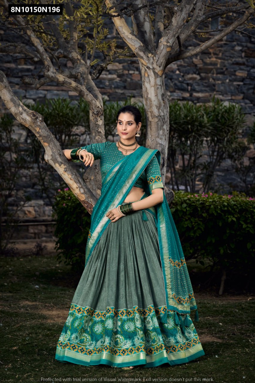 Full body shot of a woman in a stunning TRENDBUY teal green and grey Tasar silk lehenga choli set, featuring floral and foil prints, posing elegantly next to a tree in a garden.