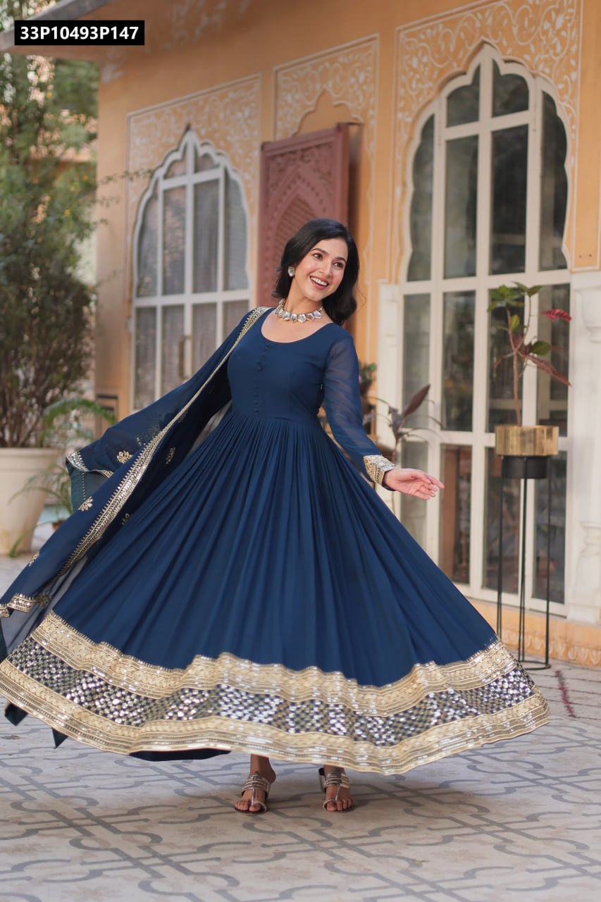 Full-body shot of a smiling model twirling in a deep teal faux blooming anarkali gown with golden zari and sequin work, paired with a matching dupatta, in a traditional courtyard setting.