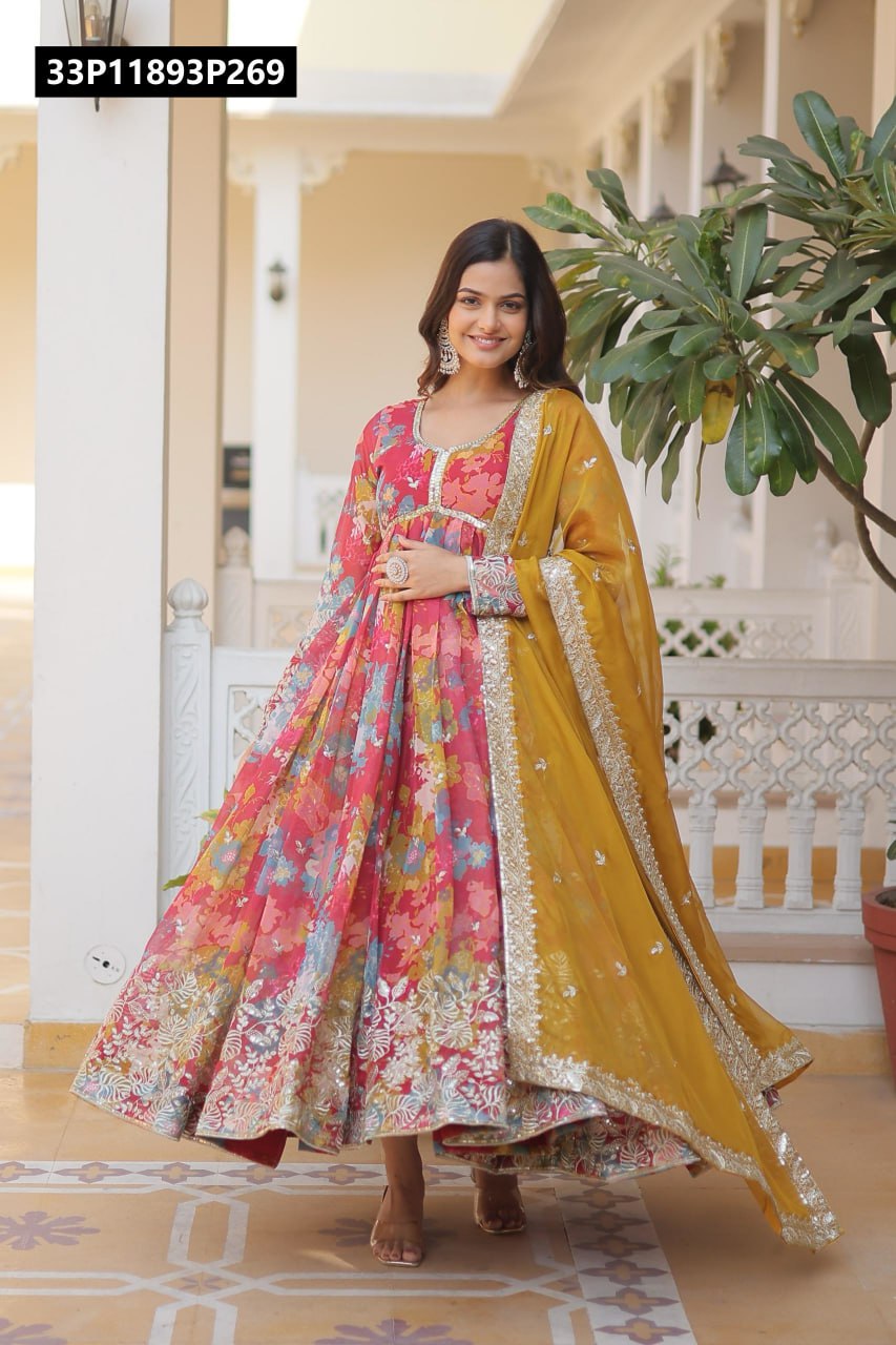 Full body shot of a woman smiling, wearing a vibrant pink-red multi-color floral digital printed Russian silk Anarkali gown with a mustard yellow embroidered dupatta, standing outdoors.