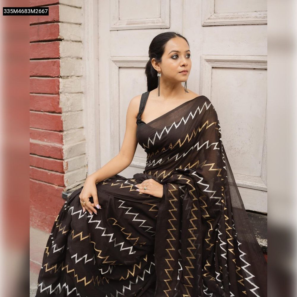 Model sitting on steps, wearing a black Fox Georgette saree with white and golden zigzag thread work, against a white door and red brick wall.