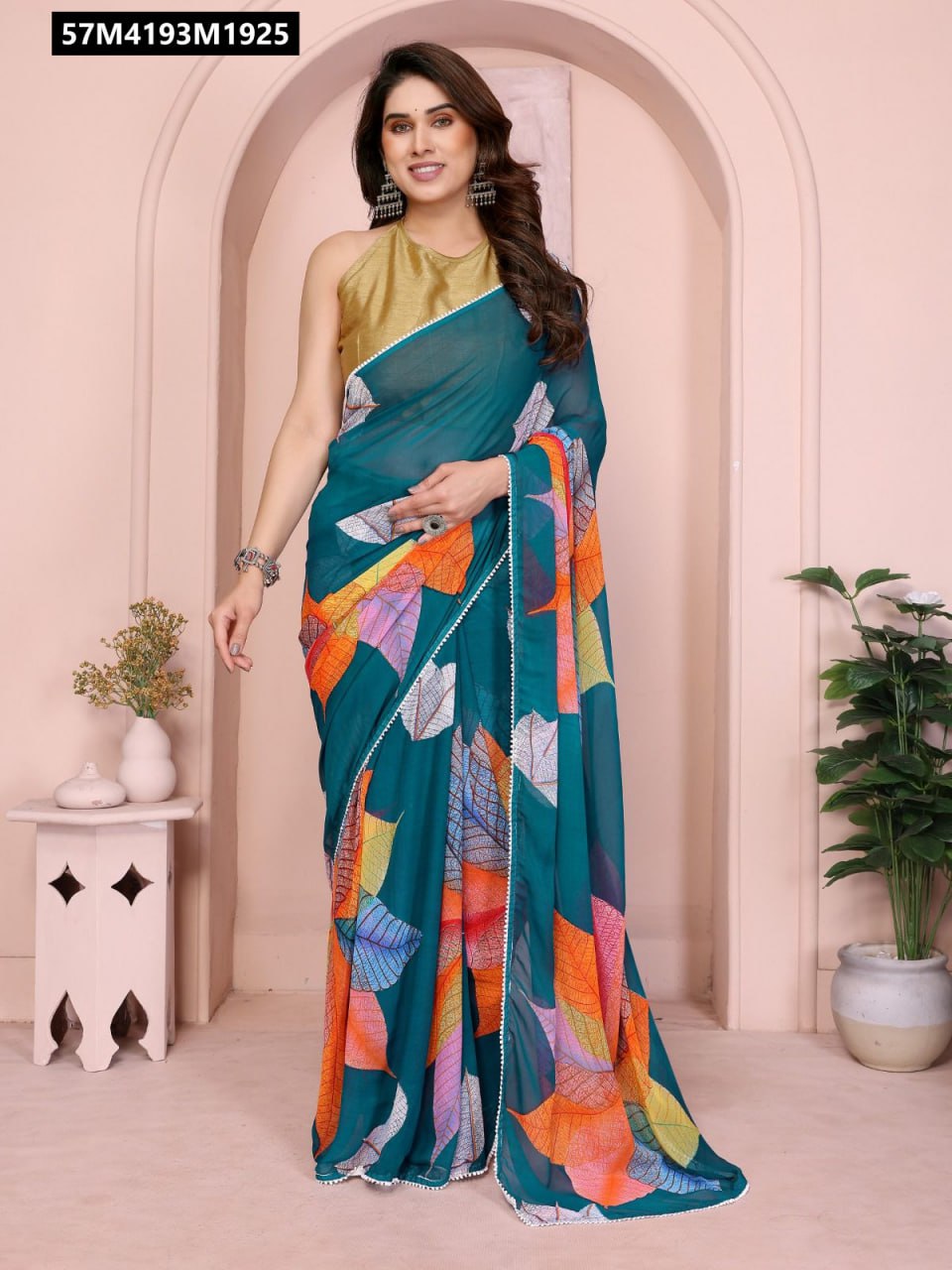 Full shot of a model wearing a teal georgette saree with vibrant multi-color leaf print and white lace border, posing against a light pink wall.