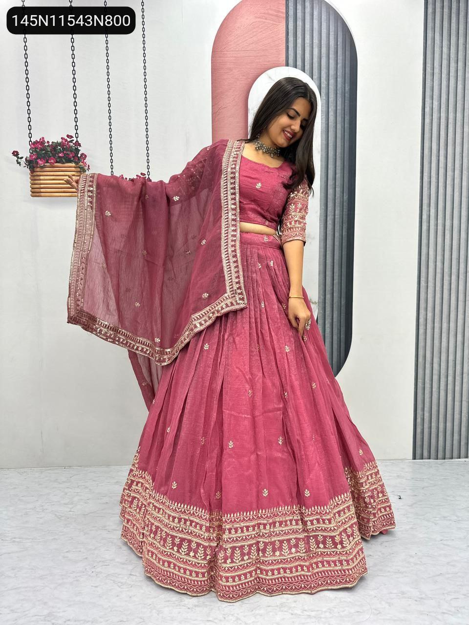 Model smiling in a pink Pure Gold Crush lehenga choli, featuring heavy gold embroidery and sequin work, with matching dupatta draped over her arm, against a modern white and pink background. (Main Image)