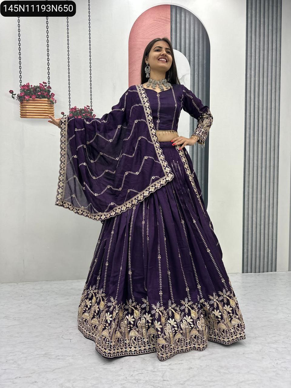 Full shot of a smiling model in a royal purple Vichitra silk lehenga choli with intricate gold thread and sequence embroidery, posing against a white and patterned wall.