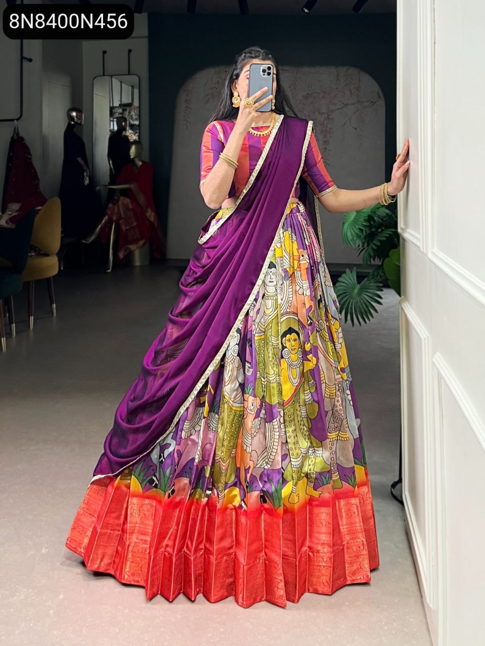 Full body shot of a woman wearing a TRENDBUY Kalamkari print Dola silk lehenga choli with an orange-red border, purple dupatta, and striped blouse, posing in an indoor setting.
