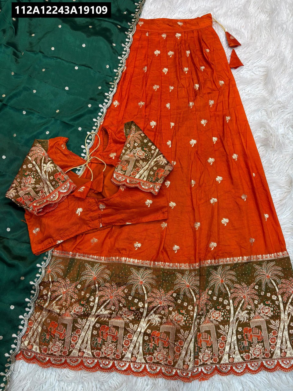 Flat lay of TRENDBUY Orange Kanchipuram Jacquard Lehenga Choli set with dark green contrast dupatta, featuring elephant and palm tree border.