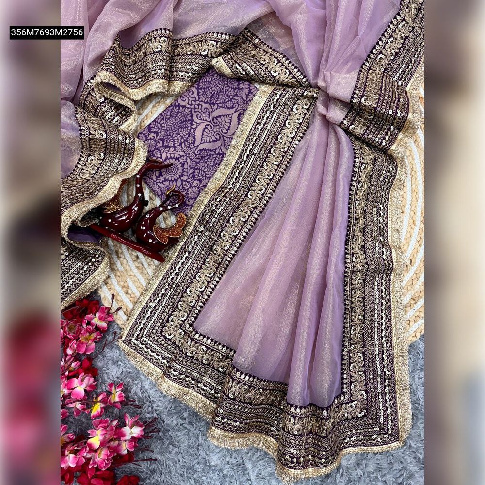 TRENDBUY lavender soft twill net saree with sequin and Dori embroidered border, laid flat with a purple jacquard blouse piece, decorative items, and pink flowers on a textured mat.