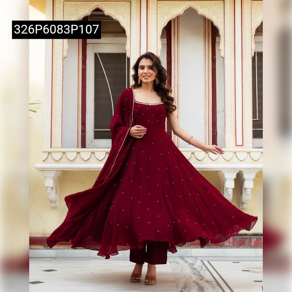 Smiling model twirling in a stunning deep red TRENDBUY Mugdha Anarkali suit, highlighting the expansive flair adorned with gold sequin embellishments and the flowing dupatta, set against a traditional backdrop.