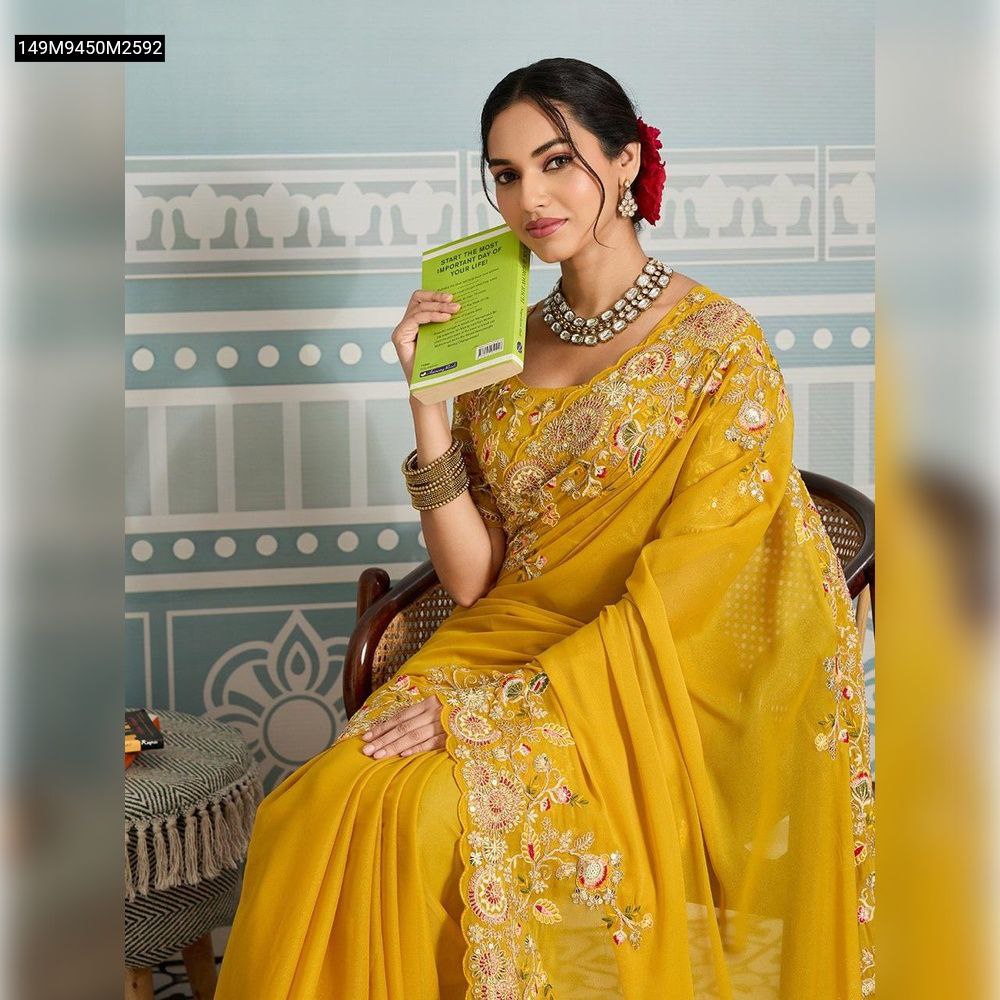 Close-up of a model wearing the TRENDBUY Mustard Yellow Soft Georgette Saree, sitting and holding a book, showcasing the intricate embroidery on the pallu and the matching blouse detail.