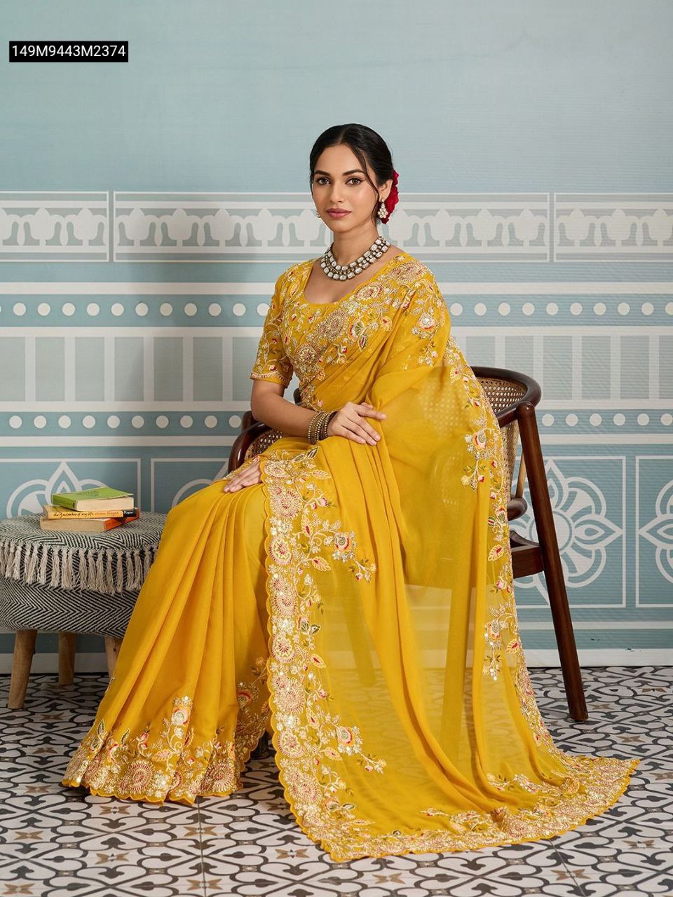 Elegant model seated, gracefully draped in the TRENDBUY Mustard Yellow Soft Star Georgette Saree, showcasing the full embroidery on the pallu and border, with a traditional backdrop.