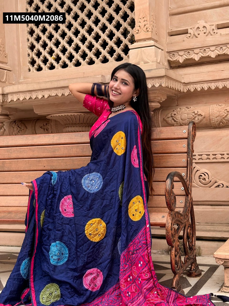 Young woman smiling and posing on a wooden bench, wearing a TRENDBUY Navy Blue Pure Moss Bandhani Saree with vibrant circular mirror work and a bright pink bandhani print pallu, showcasing the elegance of the drape.