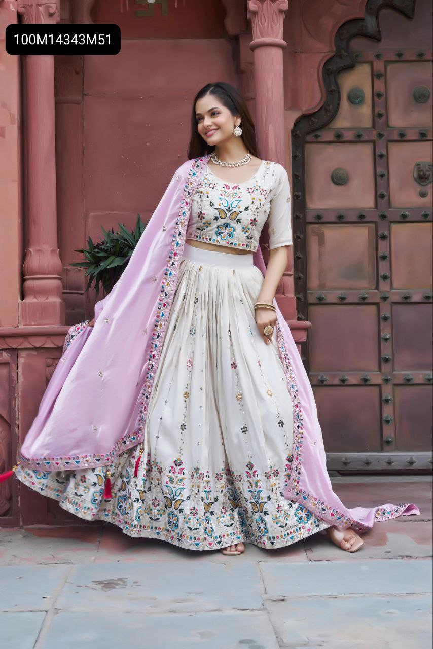 A female model in an off-white tissue silk lehenga choli with a light lavender dupatta, standing elegantly against a traditional reddish-pink door, smiling softly.