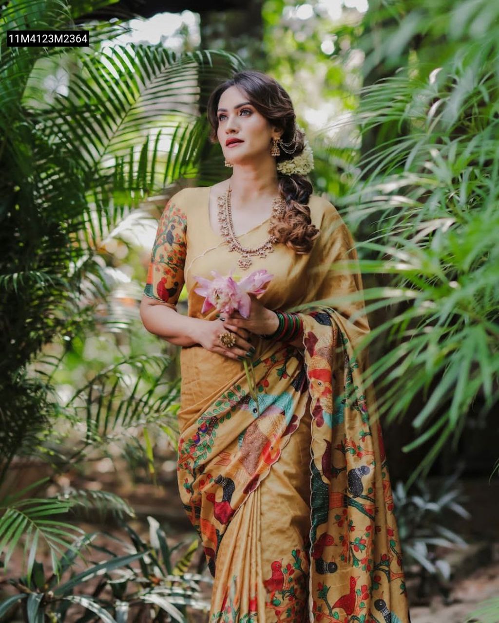 A smiling woman gracefully posing amidst lush green foliage, wearing a TRENDBUY Mustard Handloom Tussar Silk Saree with a vibrant Kalamkari bird and tree digital print, holding a pink lotus flower.