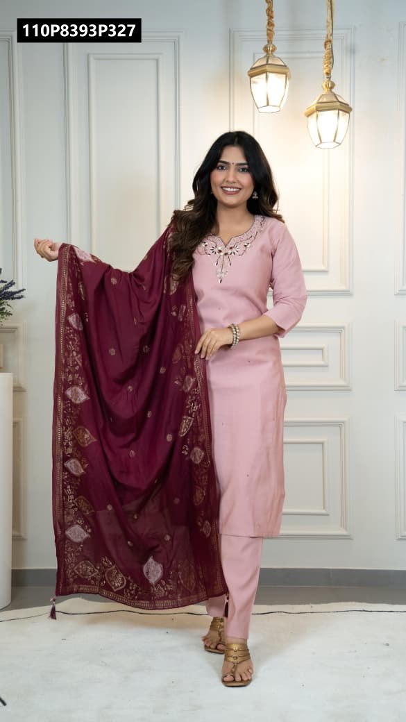 Full shot of a smiling model in a light pink Roman Silk kurta set with a maroon jacquard dupatta, posing elegantly against a white panelled wall.