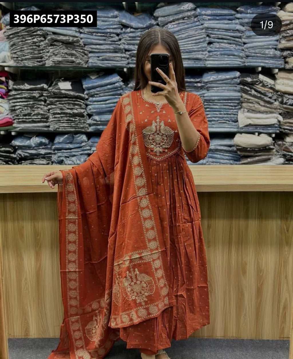 Model wearing a vibrant rust orange pure cotton Anarkali kurta set with rich golden embroidery and a patterned mulmul dupatta, against a store backdrop.