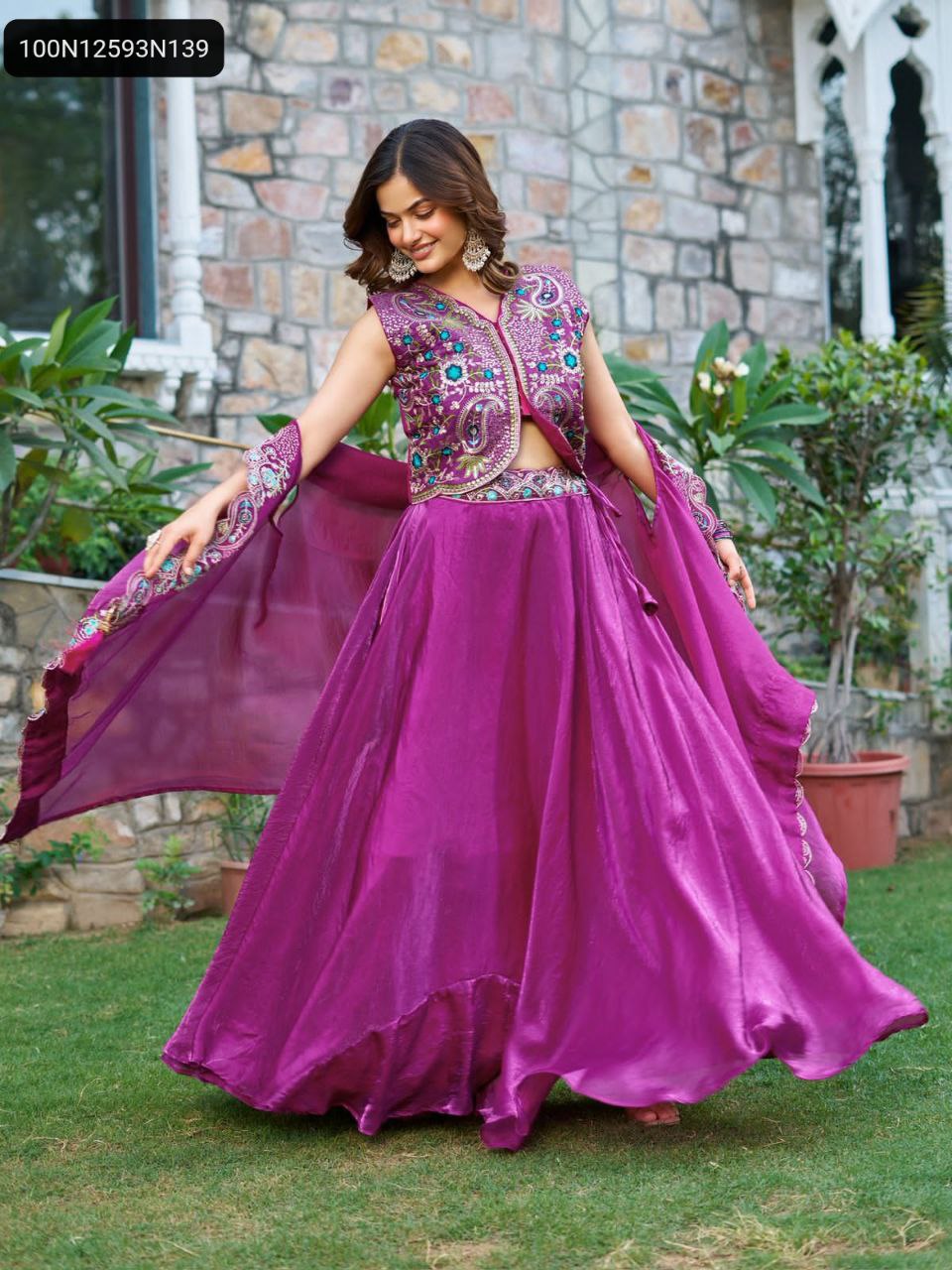 Full body shot of a smiling model in a magenta Fendy Silk Lehenga Choli with intricate embroidery and a flowing shrug dupatta, twirling on green grass in front of a stone wall.