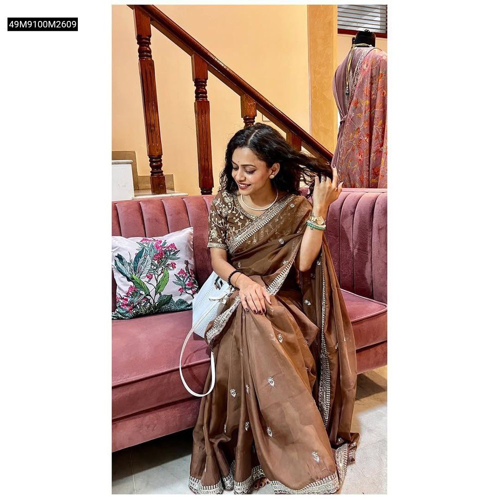 Model showcasing the TRENDBUY Brown Pure Soft Organza Saree, elegantly draped, featuring heavy zari and sequence work, while seated gracefully on a pink sofa in a well-lit living space.