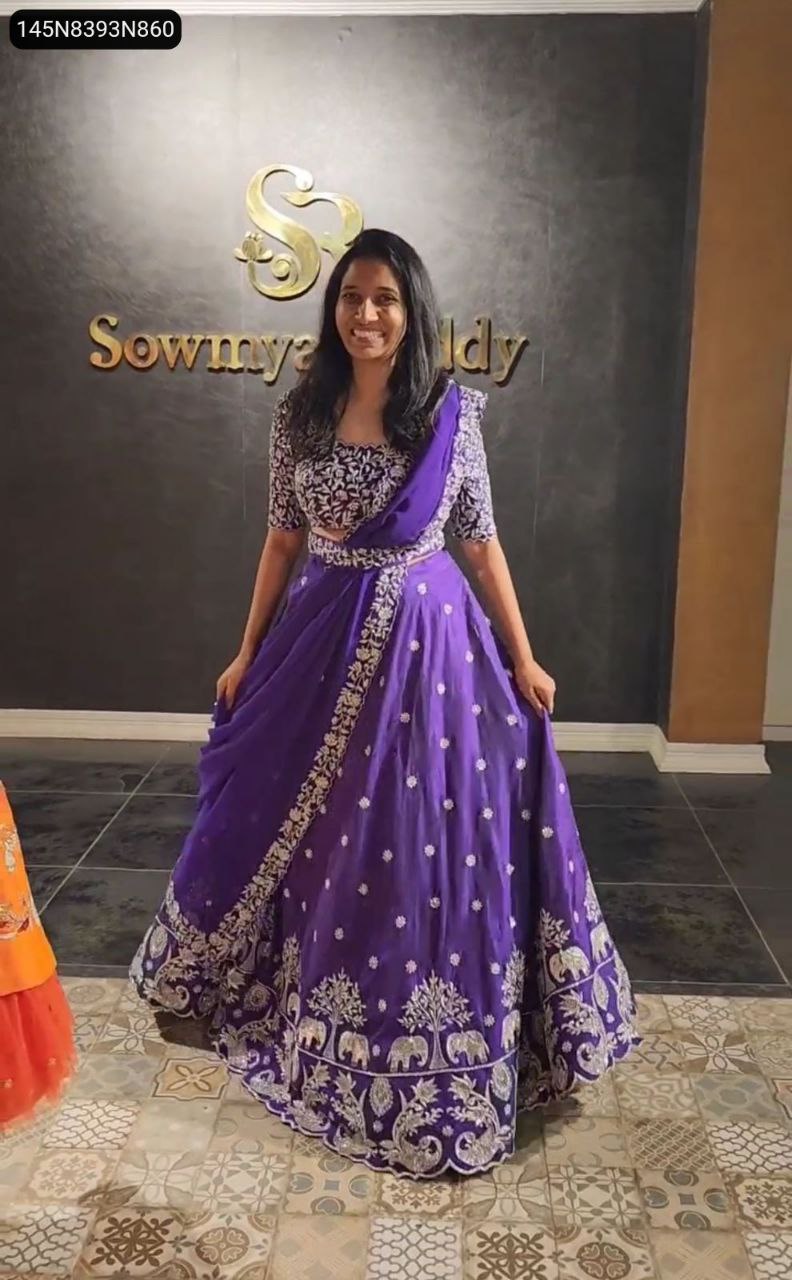 Full shot of a smiling model in a vibrant purple embroidered Chinon lehenga choli with dupatta, standing elegantly against a dark backdrop.
