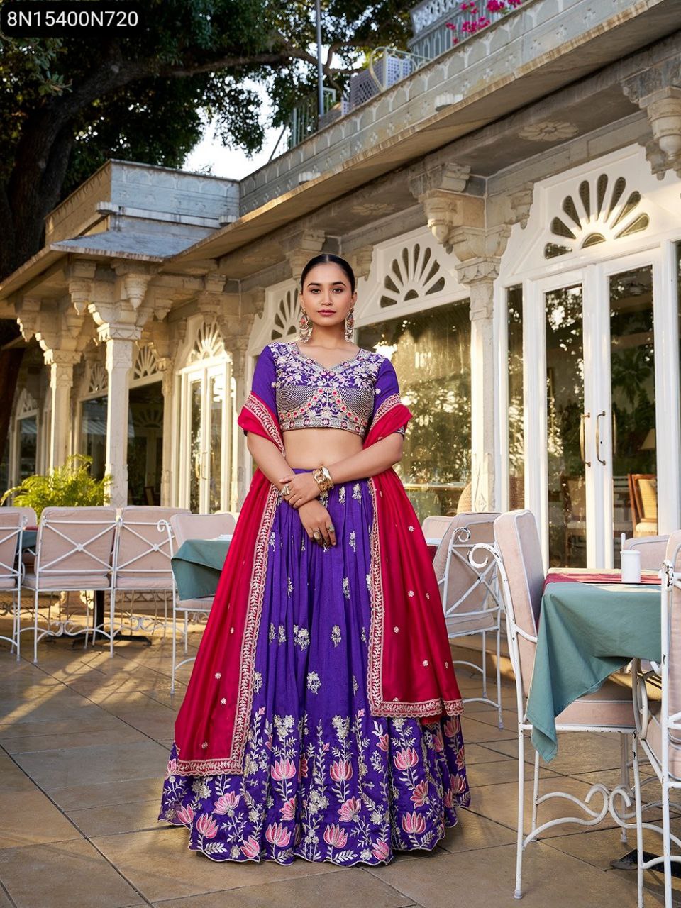 Full body shot of model elegantly posing in TRENDBUY purple and magenta embroidered Vichitra silk lehenga choli, featuring floral patterns and a wide flair, set against an outdoor palace-like backdrop.