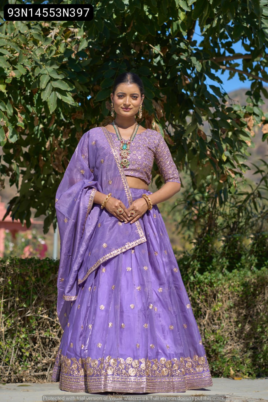 Full body shot of a model elegantly posing in a radiant purple Gold Crush lehenga choli, featuring detailed gold embroidery on the skirt hem and blouse, against a lush green outdoor background.