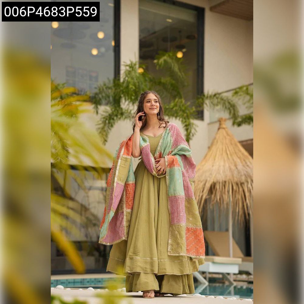Full body shot of a woman smiling, wearing a TRENDBUY Olive Green shimmer Anarkali gown with a vibrant patchwork dupatta, standing by a pool in an outdoor setting.