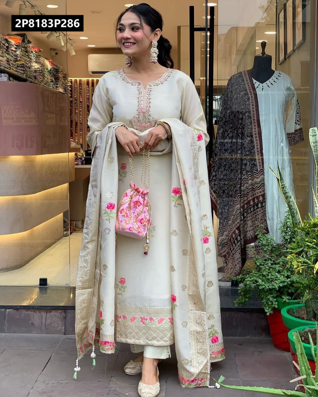 Model in cream shimmer jacquard kurta set with detailed pink and gold embroidery, holding a potli bag, smiling in front of a store.