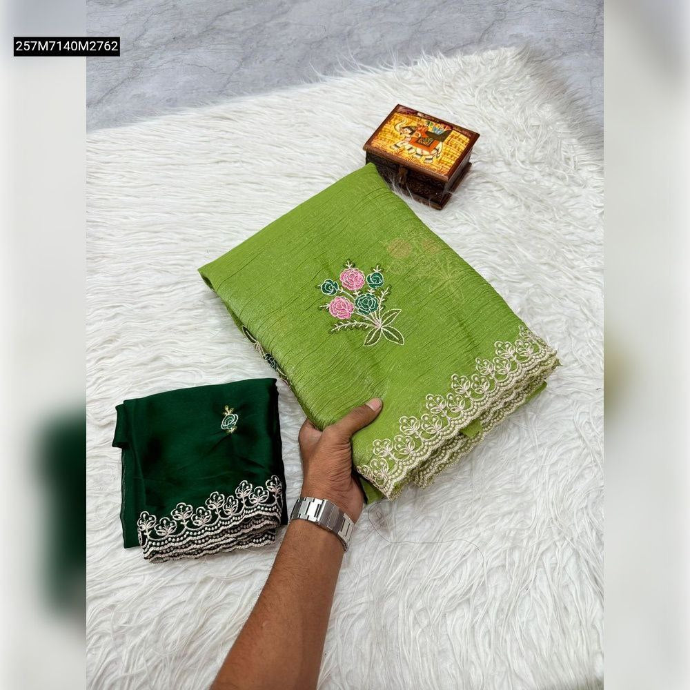 A hand holding a folded vibrant green TRENDBUY soft crunchy silk saree with delicate multi-color floral embroidery and a white lace border, alongside a folded dark green contrast Rangoli silk blouse piece, displayed on a plush white surface.